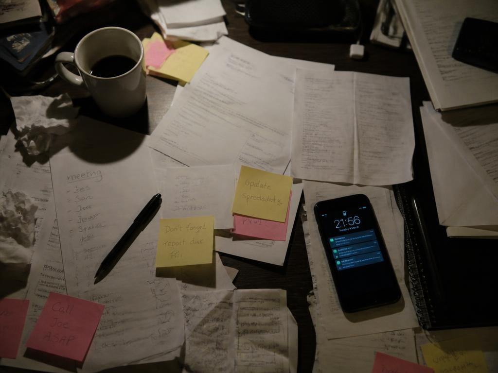 A cluttered desk covered in papers and sticky notes next to a phone full of notifications